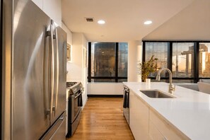 A modern kitchen with a stainless steel refrigerator, a stove, and a large window providing a view of the city. - Skyline V | 26th-floor 3BR w/Doorman, Gym, and Pool (New York)