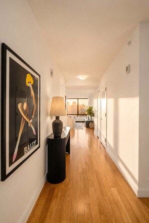 A hallway with wooden flooring, a framed artwork on the wall, a lamp on a side table, and a view of the city through a window. - Skyline V | 26th-floor 3BR w/Doorman, Gym, and Pool (New York)