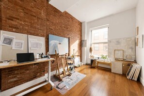 A room with a brick wall, wooden floor, easels, a laptop, and artwork on the walls. - Greene Loft by StayHaus | 3BR w/ Doorman, Elevator (New York)
