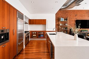 A modern kitchen with wooden cabinets, stainless steel appliances, a brick wall, and a large island with a sink. - Greene Loft by StayHaus | 3BR w/ Doorman, Elevator (New York)