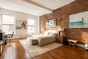 A bedroom with a brick wall, a bed, a desk, and a chair. - Greene Loft by StayHaus | 3BR w/ Doorman, Elevator (New York)