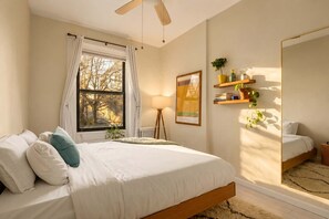 A bedroom with a bed, a window with curtains, a bedside lamp, a bedside plant, a wall-mounted shelf with books and a plant, and a mirror. - Clinton Duplex by StayHaus | 2BR with Private Yard (Brooklyn)