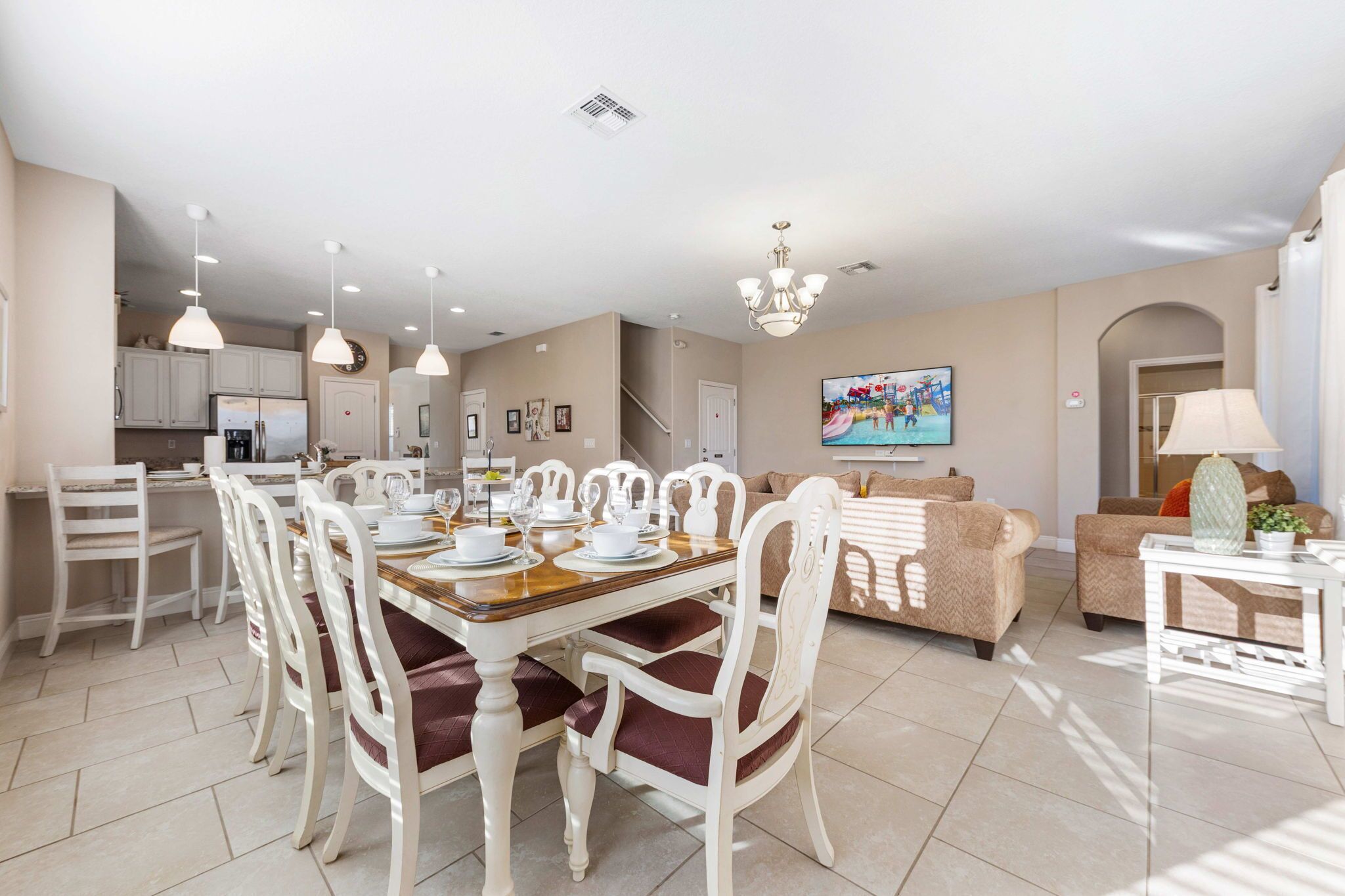 A spacious living area with a dining table set for four, surrounded by white chairs with red cushions. A beige sofa and a white armchair are positioned in the background.