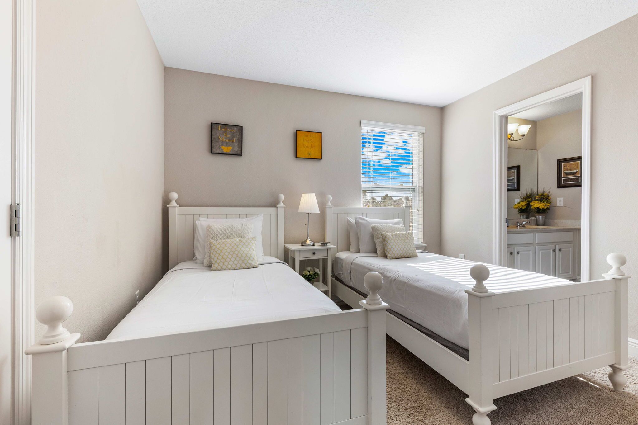 A bedroom with two white beds, a nightstand, a lamp, and a window with blinds.