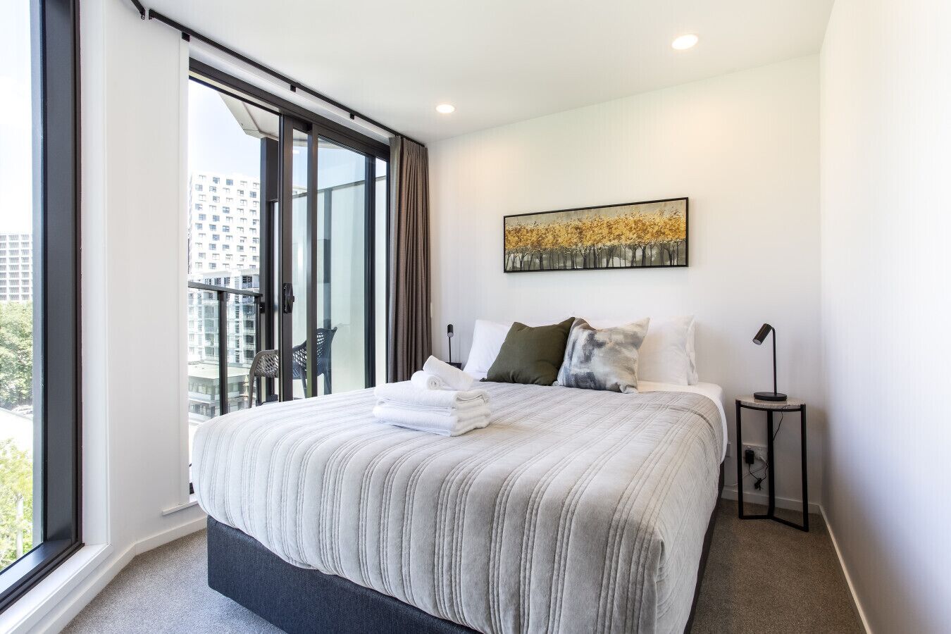 A modern hotel room with a large bed, a view of the city through floor-to-ceiling windows, a painting on the wall, and a small bedside table with a lamp.