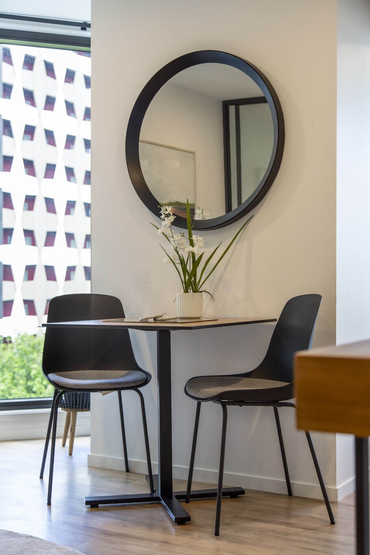 A modern dining area with two black chairs, a round mirror, and a small white vase with flowers on a table.