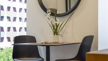 A modern dining area with two black chairs, a round mirror, and a small white vase with flowers on a table.