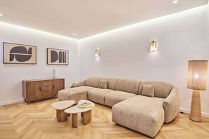 Ein modernes Wohnzimmer mit einer Eckcouch, Holzdielen im Fischgrätmuster, einer Kommode und zwei kleinen Tischen.