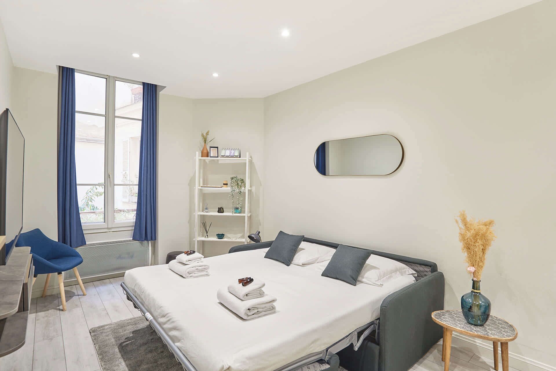 Une chambre d’hôtel moderne avec un grand lit, un bureau, une chaise, une petite table, un miroir et une fenêtre avec des rideaux.