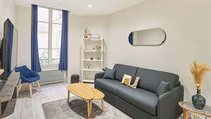 A modern living room with a grey sofa, a wooden coffee table, a blue chair, a white shelving unit, and a large window with blue curtains. - Spacious Studio Sacre Coeur-Montmartre (Paris)