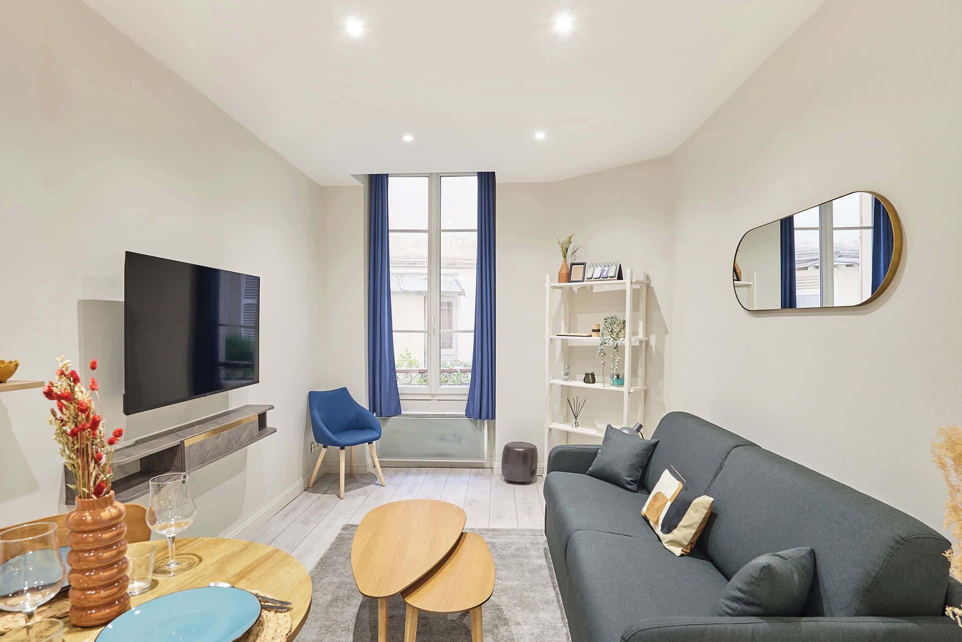 Un salon moderne avec un téléviseur à écran plat, un fauteuil bleu, un canapé gris et une étagère blanche.