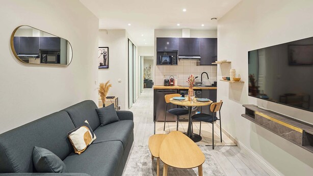 A modern living room with a dark grey sofa, a wooden coffee table, and a TV on the right. A kitchen with dark cabinets and a microwave is visible in the background. - Spacious Studio Sacre Coeur-Montmartre (Paris)