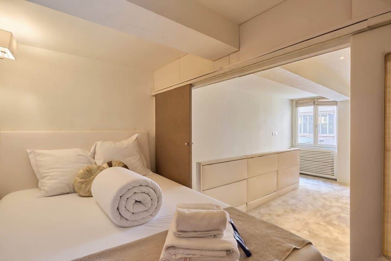 A hotel room with a bed, a bedside table with towels, a headboard, and a view of the adjacent room.