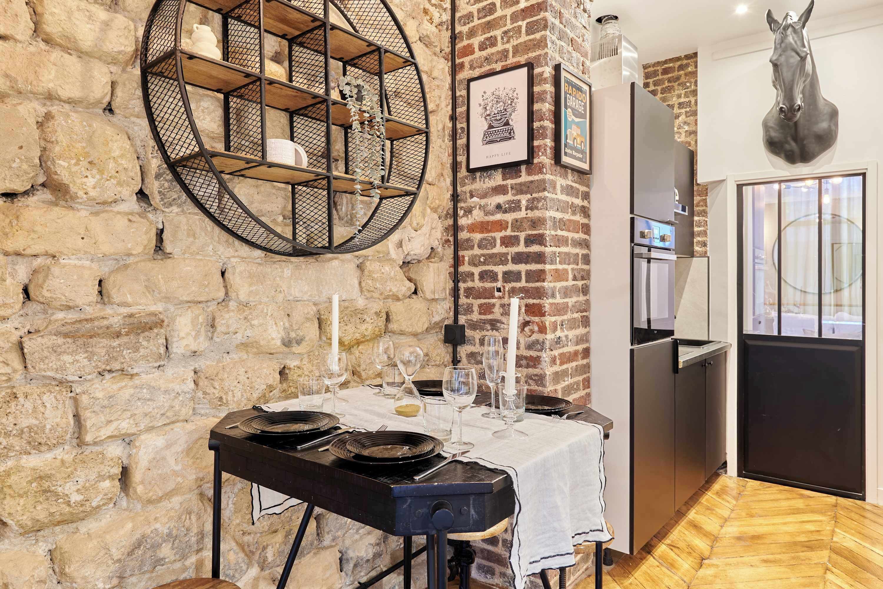 A rustic kitchen with a stone wall, a round metal shelf, a refrigerator, and a dining table set with plates and glasses.