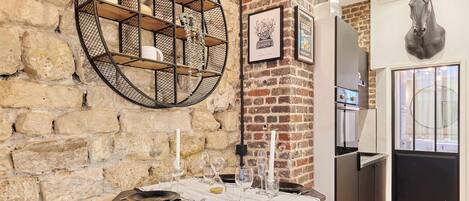 A rustic kitchen with a stone wall, a round metal shelf, a refrigerator, and a dining table set with plates and glasses.