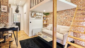 A modern living space with a brick wall, a loft area with a wooden ladder, a sofa, a coffee table, and a dining area with a table and chairs. - Charming Apartment - Paris Pere Lachaise (Paris)