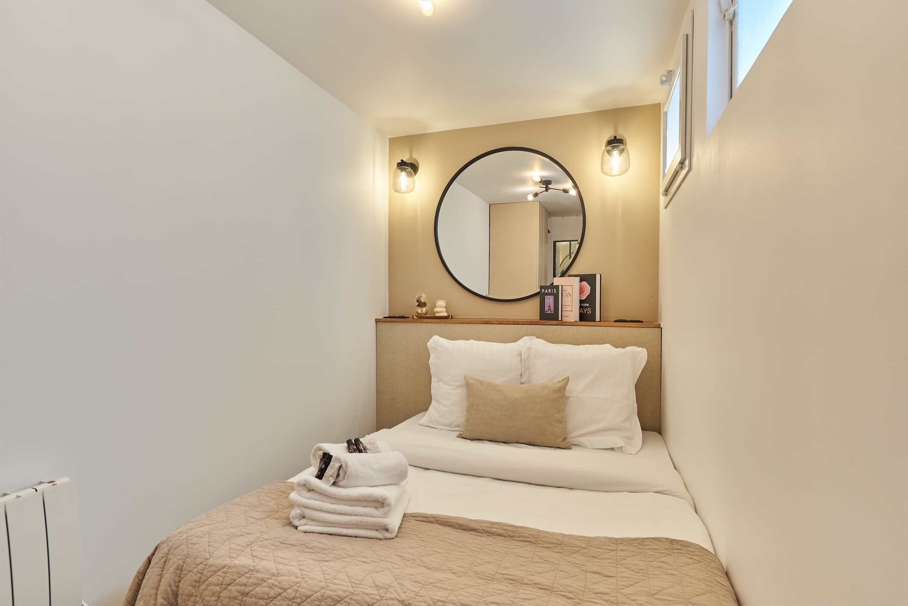 A neatly made bed with a beige quilt, a round mirror above it, and a small bedside table with a book and a lamp.