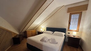 Una camera da letto con soffitto spiovente, un letto con lenzuola bianche, due comodini con lampade e una finestra con persiane.