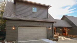 A two-story house with a garage, stone foundation, and a small porch with wicker chairs. - Sapphire Retreat • Stylish 1BR Suite + Hot Tub, Views, Ski Hill & Outdoor Living (Sapphire)