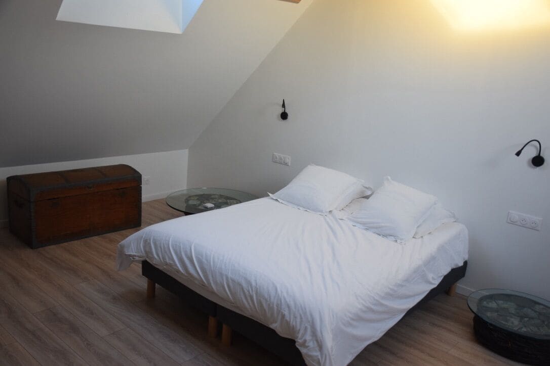 Un dormitorio con una cama, un baúl de madera, una mesa de vidrio y una lámpara de pared.