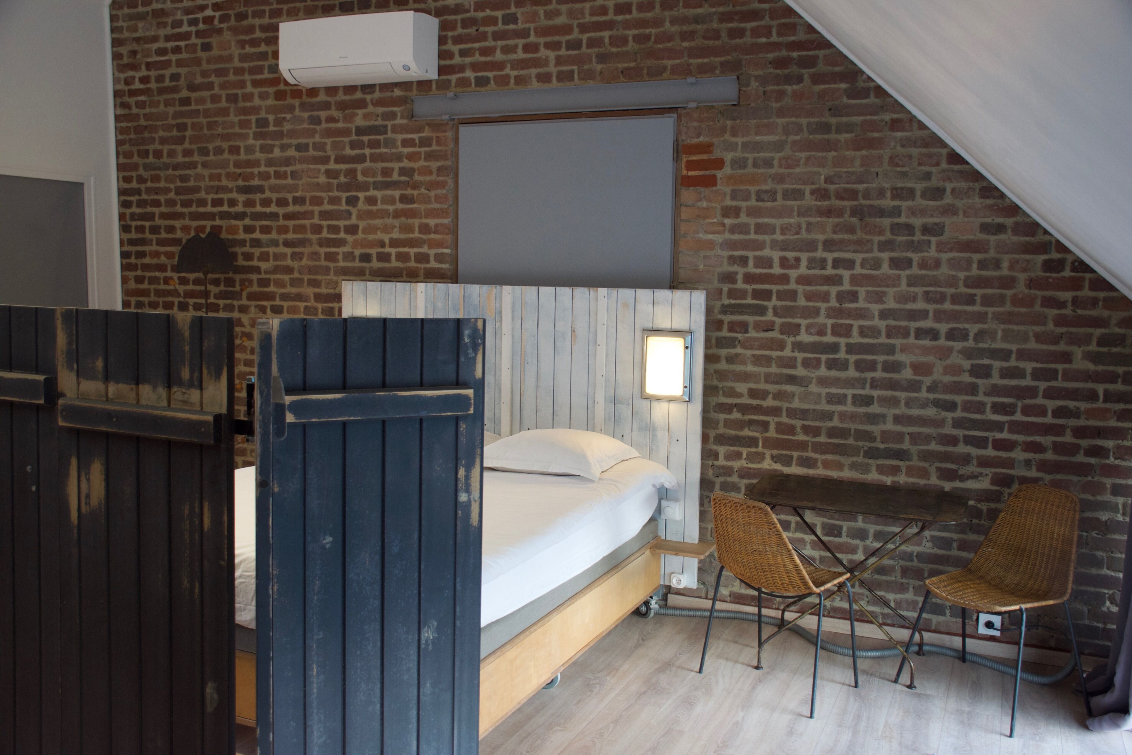 Una habitación con cama, una mesa, dos sillas, un ventanal y una pared de ladrillo.