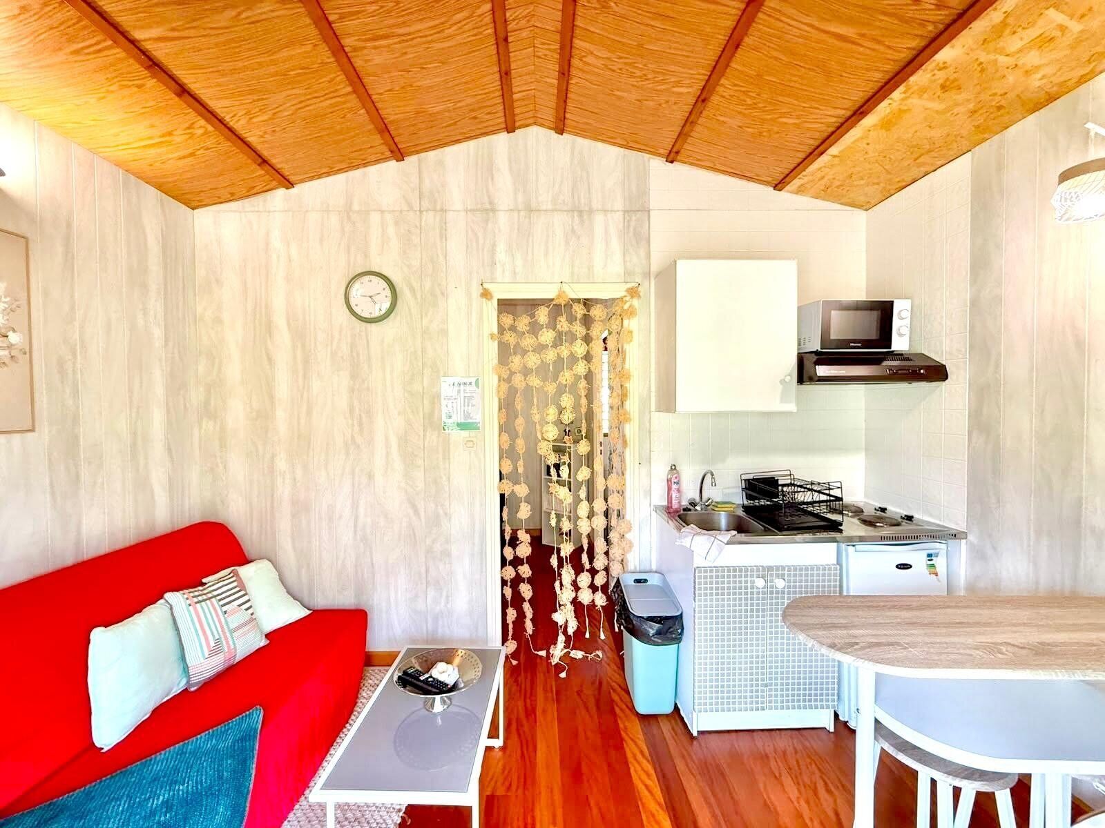 A compact living space with a kitchenette, a red sofa, a small dining table, and a wooden ceiling.