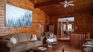 A cozy log cabin interior with a wooden ceiling, a large painting of a forest, a sofa, a chair, and a dining area with a wooden railing. - 4BR Waterfront Escape w/Woodstoves & Large Dock (Algonquin Highlands)