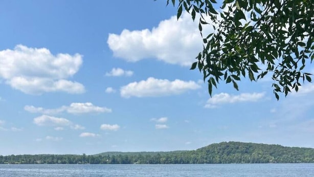 A lakeside view with a wooden dock extending into calm water, two red Adirondack chairs, and purple flowers in the foreground. - 4BR Waterfront Escape w/Woodstoves & Large Dock (Algonquin Highlands)