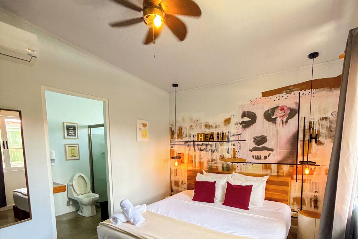 Une chambre d’hôtel avec un lit, un ventilateur de plafond, une salle de bain avec des toilettes et un lavabo, et une grande fenêtre.