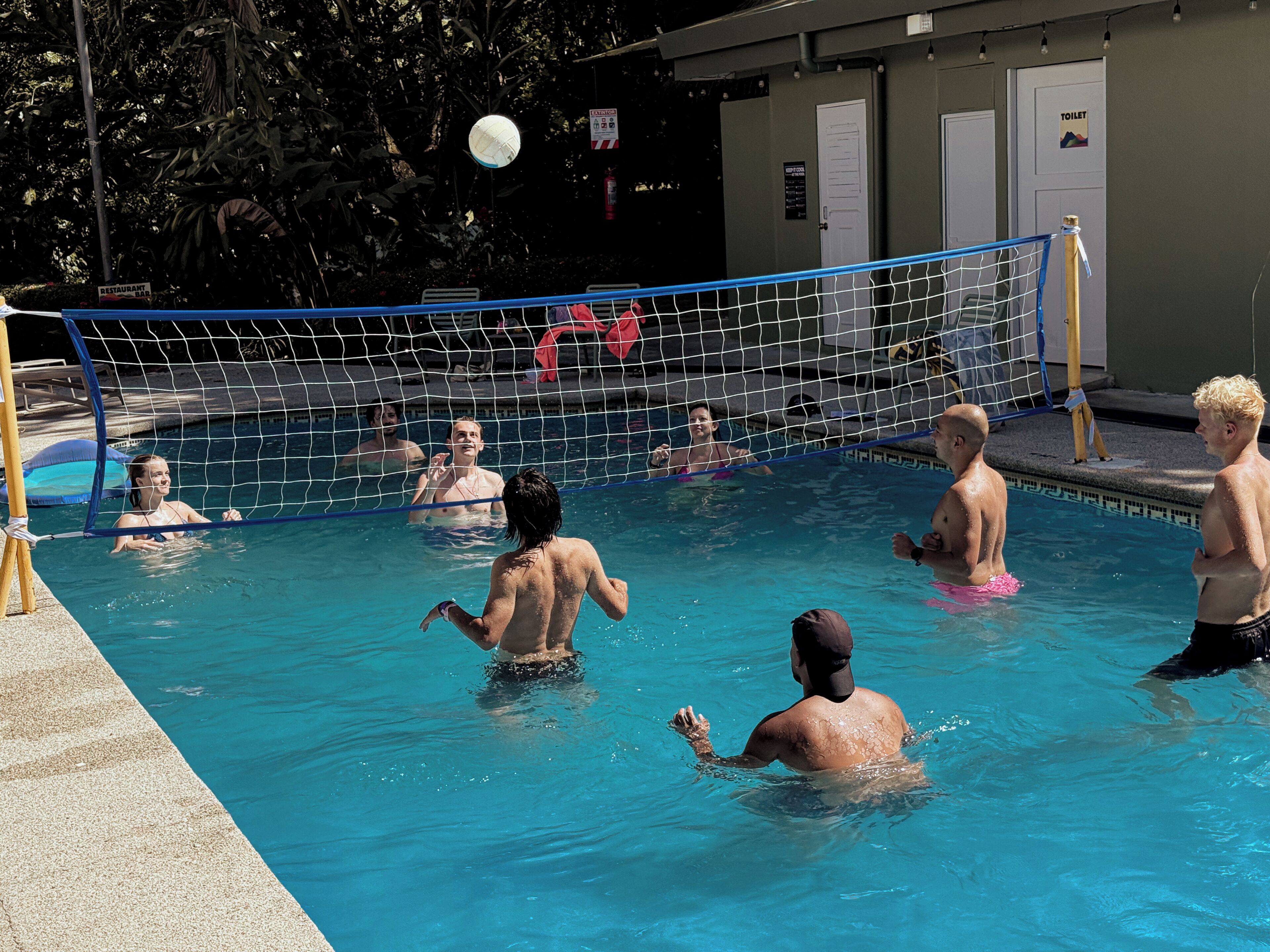 Piscina con una red de voleibol, rodeada por una zona de piscina con puertas y un letrero que indica un restaurante.