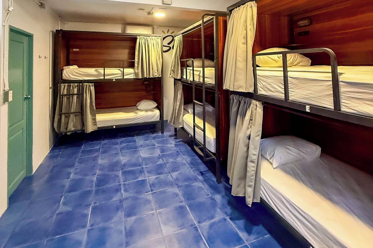 Une chambre de dortoir avec des lits superposés, un sol carrelé de carreaux bleus, une porte verte et un plafonnier blanc.