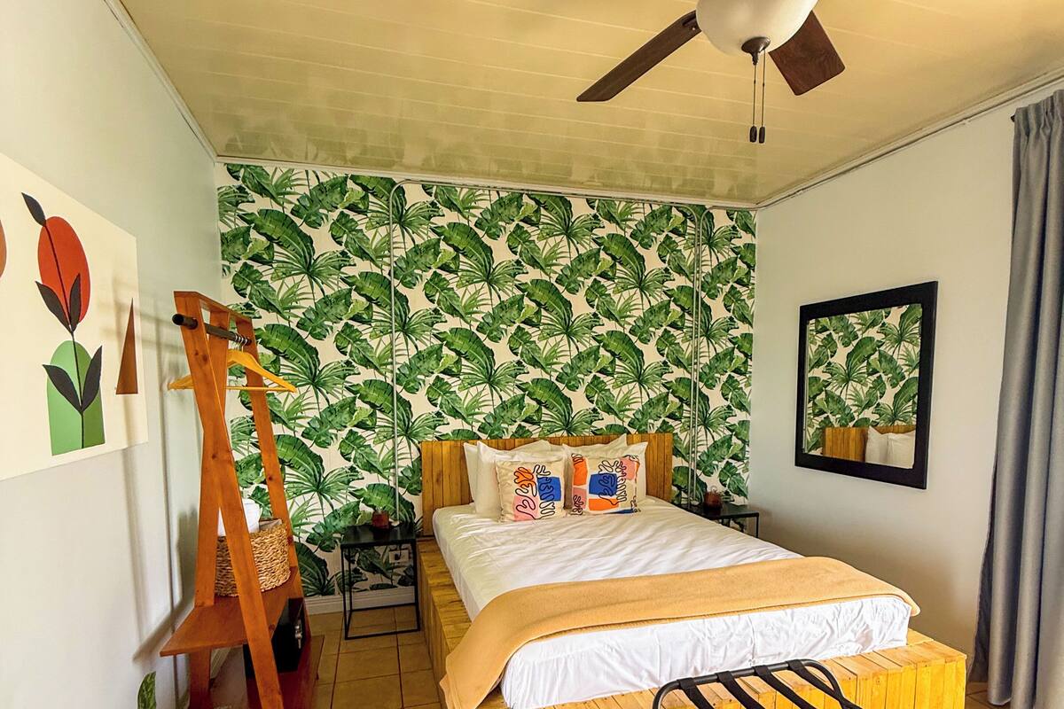 Une chambre avec un lit en bois, un ventilateur de plafond, un mur orné d’un motif végétal et une plante en pot.