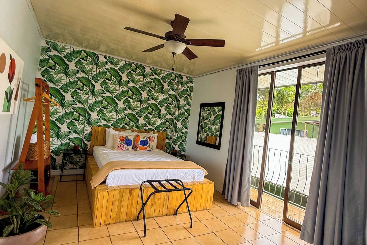 Une chambre à coucher avec un lit, un ventilateur de plafond, une grande fenêtre avec des rideaux, un banc en bois et un canapé vert.