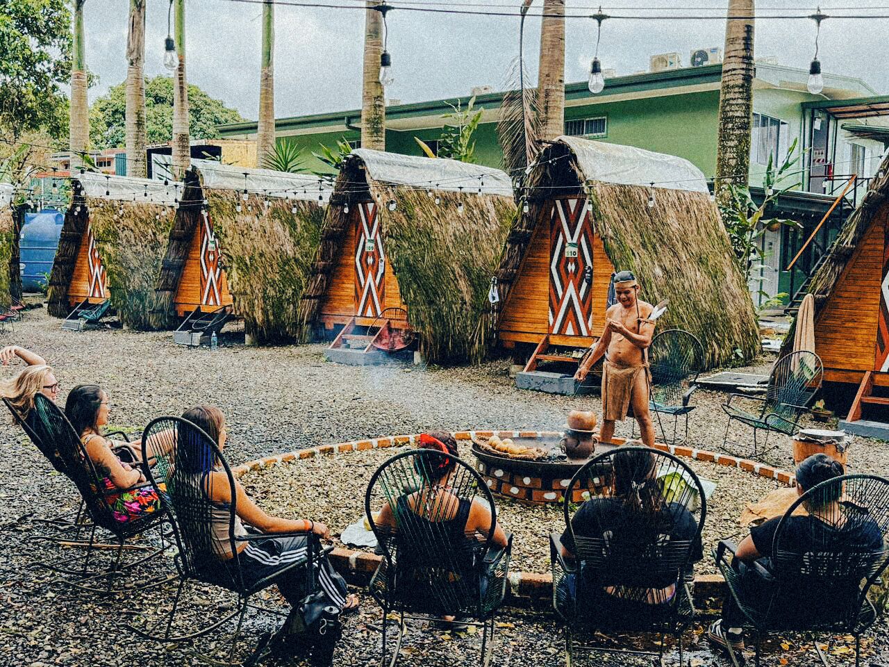 A rustic outdoor seating area with thatched-roof huts, a fire pit, and guests relaxing.