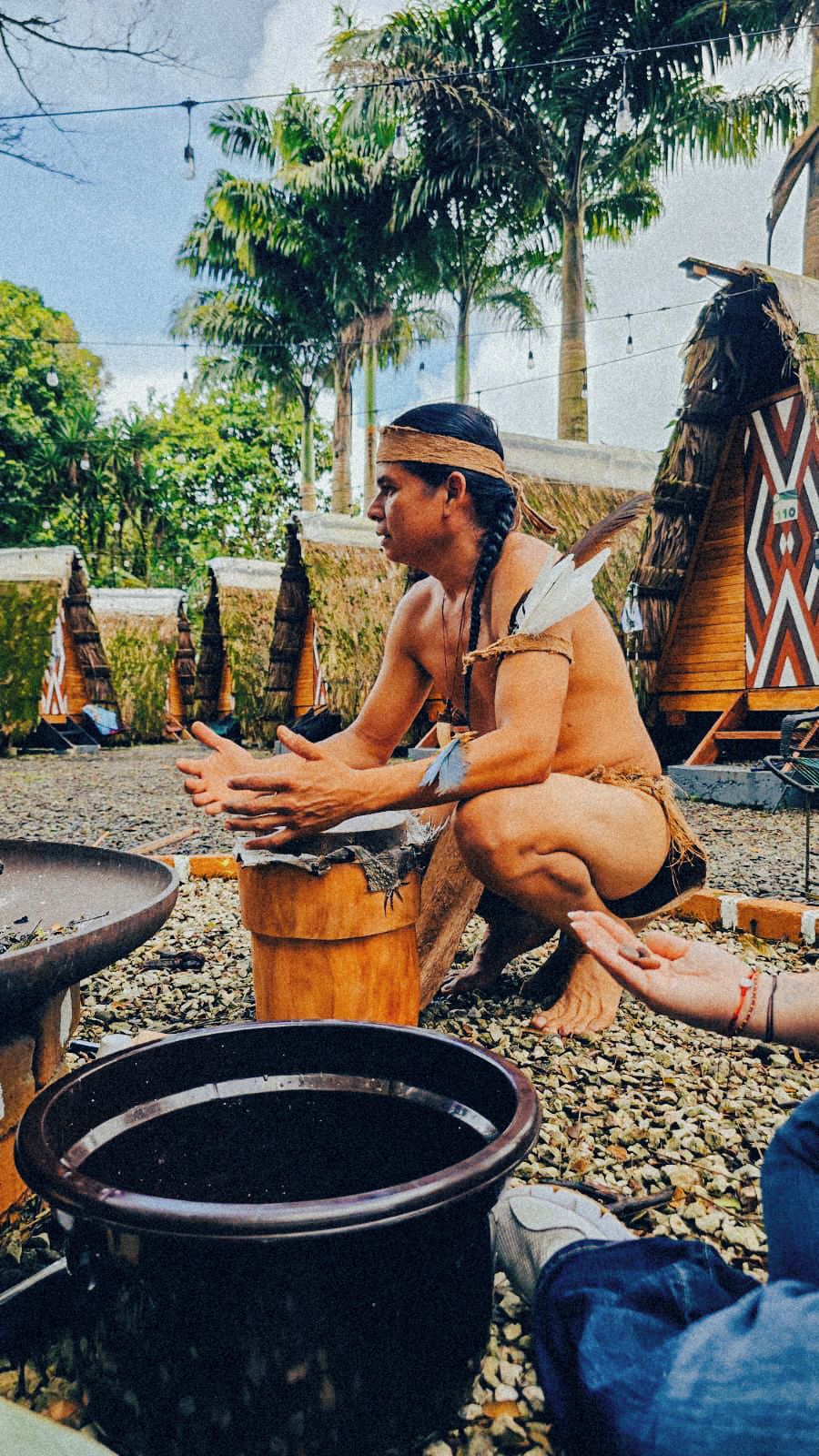 Une personne est assise sur un tabouret en bois, à l’extérieur, dans un cadre rustique. On aperçoit en arrière-plan un grand pot noir et une structure recouverte de chaume.