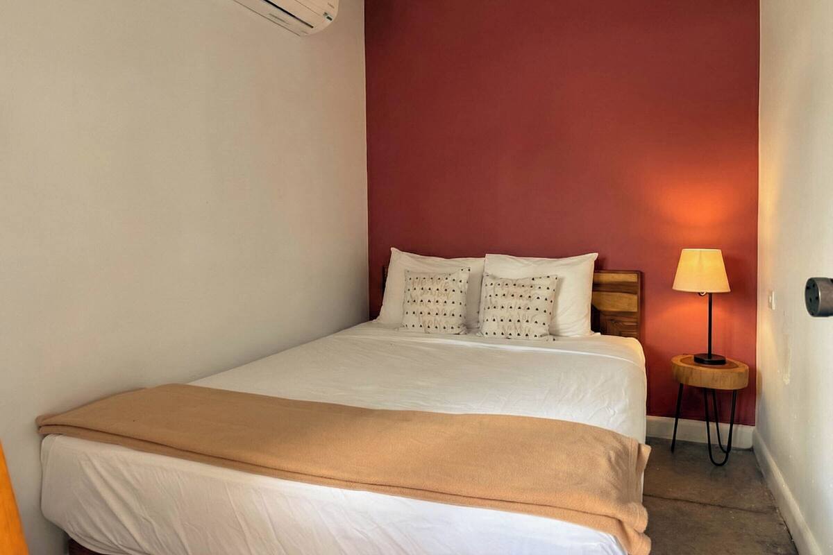 Une chambre d’hôtel avec un lit, un mur d’accent rouge, une petite table de chevet en bois avec une lampe et un appareil de climatisation.