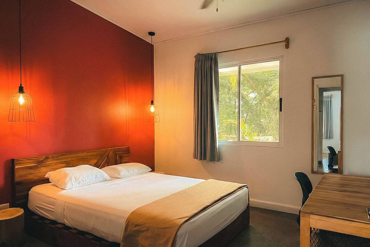 Une chambre d’hôtel avec un lit, une tête de lit en bois, une fenêtre avec des rideaux, un bureau et une chaise.