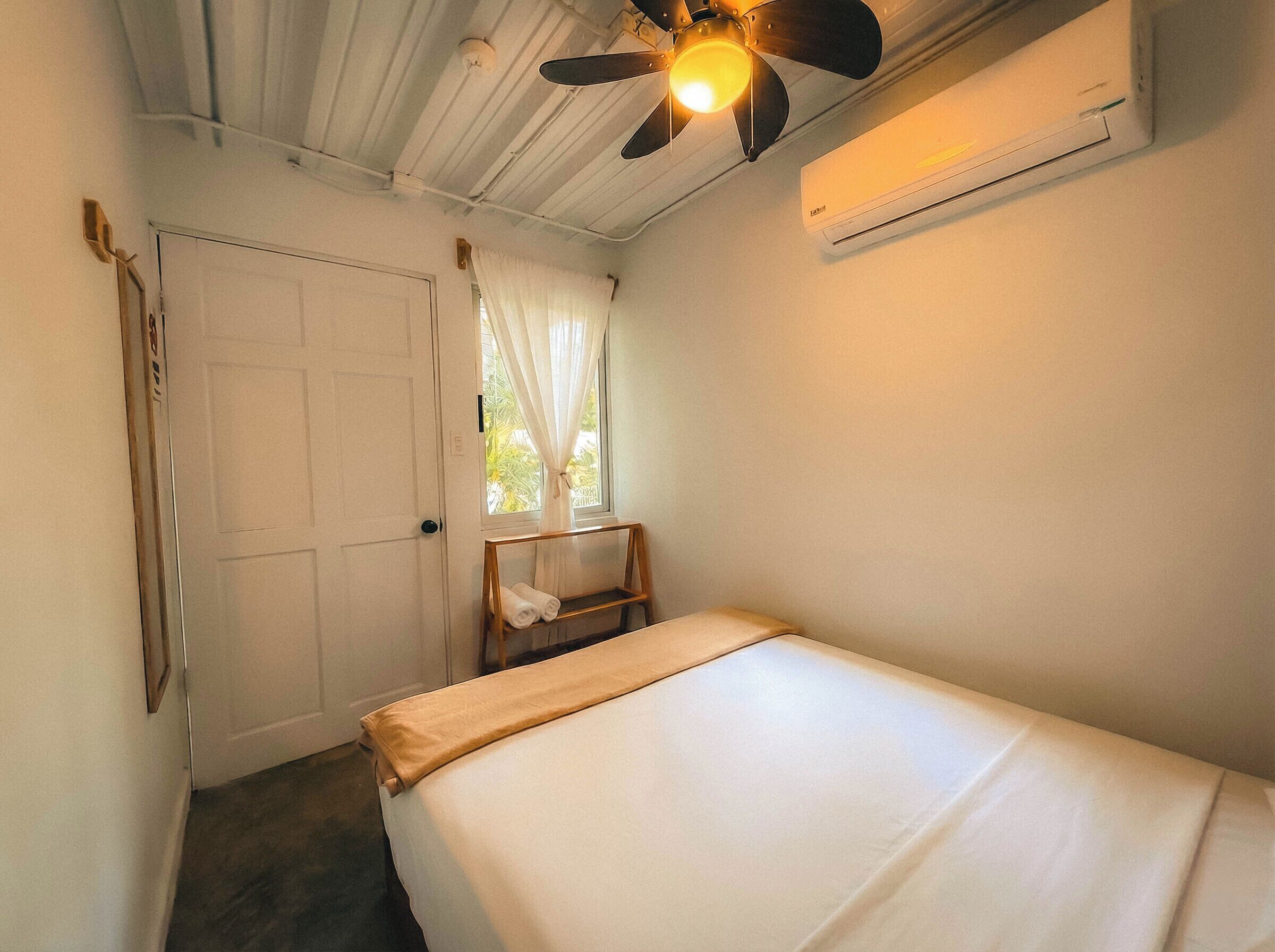 Une petite chambre avec un lit simple, un ventilateur de plafond, un appareil de climatisation, une fenêtre avec des rideaux et une chaise.