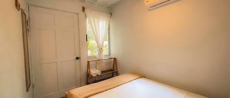 Une petite chambre avec un lit simple, un ventilateur de plafond, un appareil de climatisation, une fenêtre avec des rideaux et une chaise.