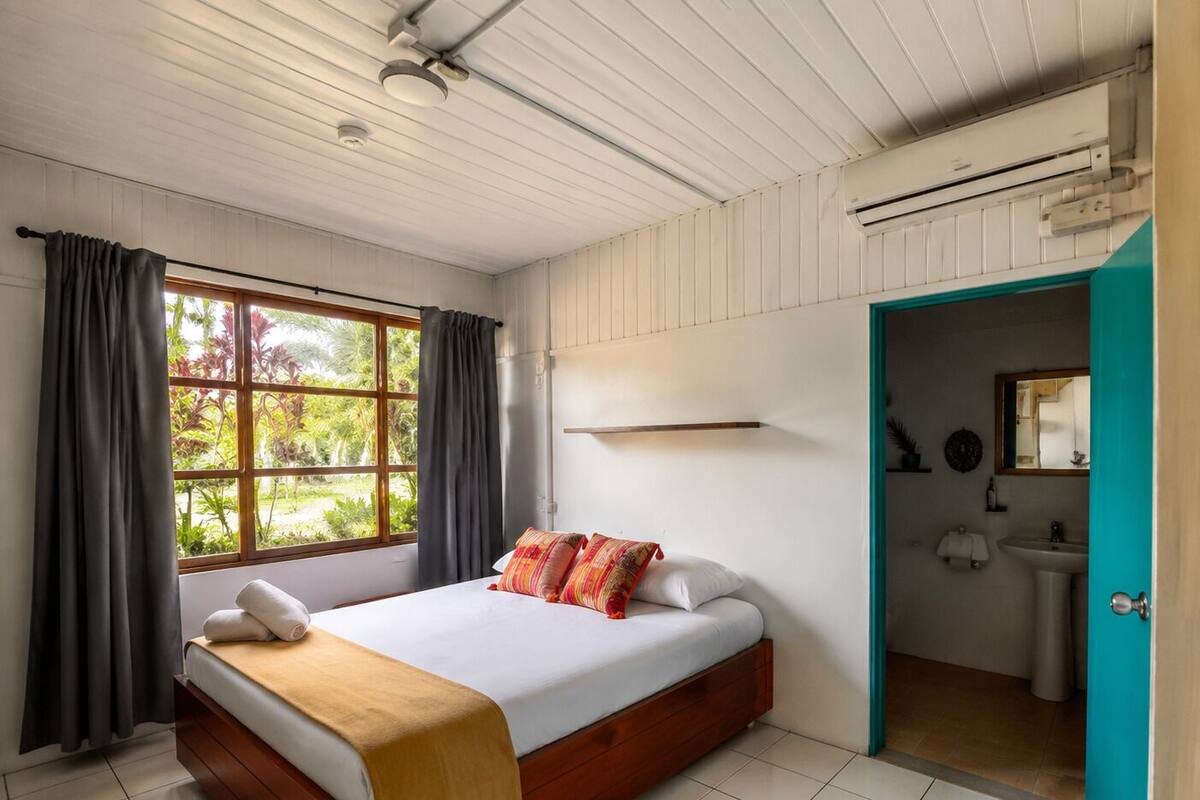 Une chambre à coucher avec un lit, une fenêtre avec des rideaux, une salle de bain avec un lavabo et un miroir, et une porte de couleur turquoise.