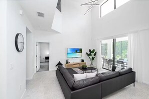 Ein modernes Wohnzimmer mit einer schwarzen Couch, einem Flachbildfernseher, einem Couchtisch und einem großen Fenster mit Blick ins Freie.