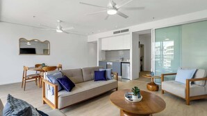 A modern living room with a sofa, chairs, a coffee table, and a dining area. There is a kitchen in the background with white cabinets and a dishwasher. - Marina Vista—Waterfront Stay near Dining & Markets (Cairns)