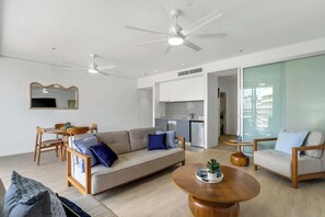 Ein modernes Wohnzimmer mit einer Couch, Sesseln, einem Couchtisch und einem Essbereich. Im Hintergrund befindet sich eine Küche mit weißen Schränken und einem Geschirrspüler.