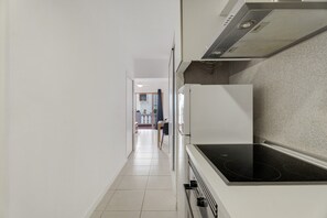 Un couloir étroit qui mène à une cuisine équipée d’une cuisinière et d’un réfrigérateur, et où l’on aperçoit, au fond, un coin repas.