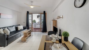 Ein modernes Wohnzimmer mit einer schwarzen Ledersofa, einem Holztisch, einem Esstisch für zwei Personen, einer Wanduhr und Blick durch eine Glastür auf einen Balkon.