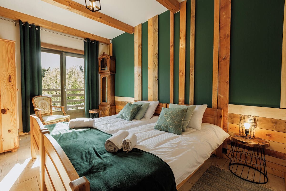 A wooden-framed bedroom with a large bed, two pillows, a nightstand, and a window with curtains.