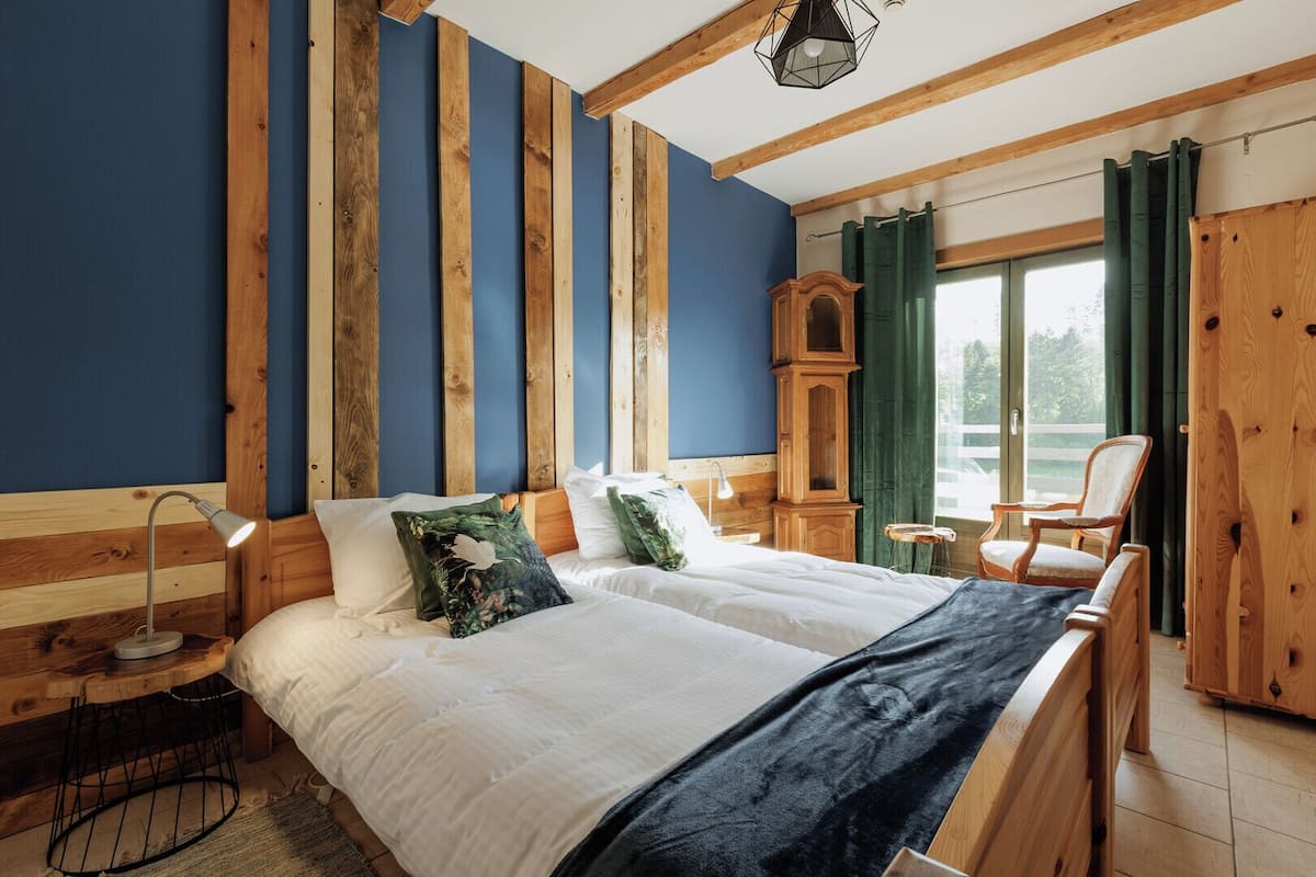 A bedroom with a large bed, wooden headboard, a desk with a lamp, a chair, and a view of the outdoors through a window.
