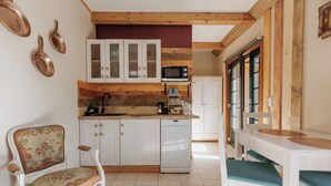 A compact kitchen with white cabinets, a microwave, and a sink. There is a dining table with chairs, and a decorative chair with floral upholstery. - Jesteśmy Na Ty Apartamenty Lewin Kłodzki (gmina Lewin Kłodzki)