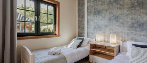 A cozy bedroom with two single beds, a nightstand with a lamp, and a window with a view of a house.