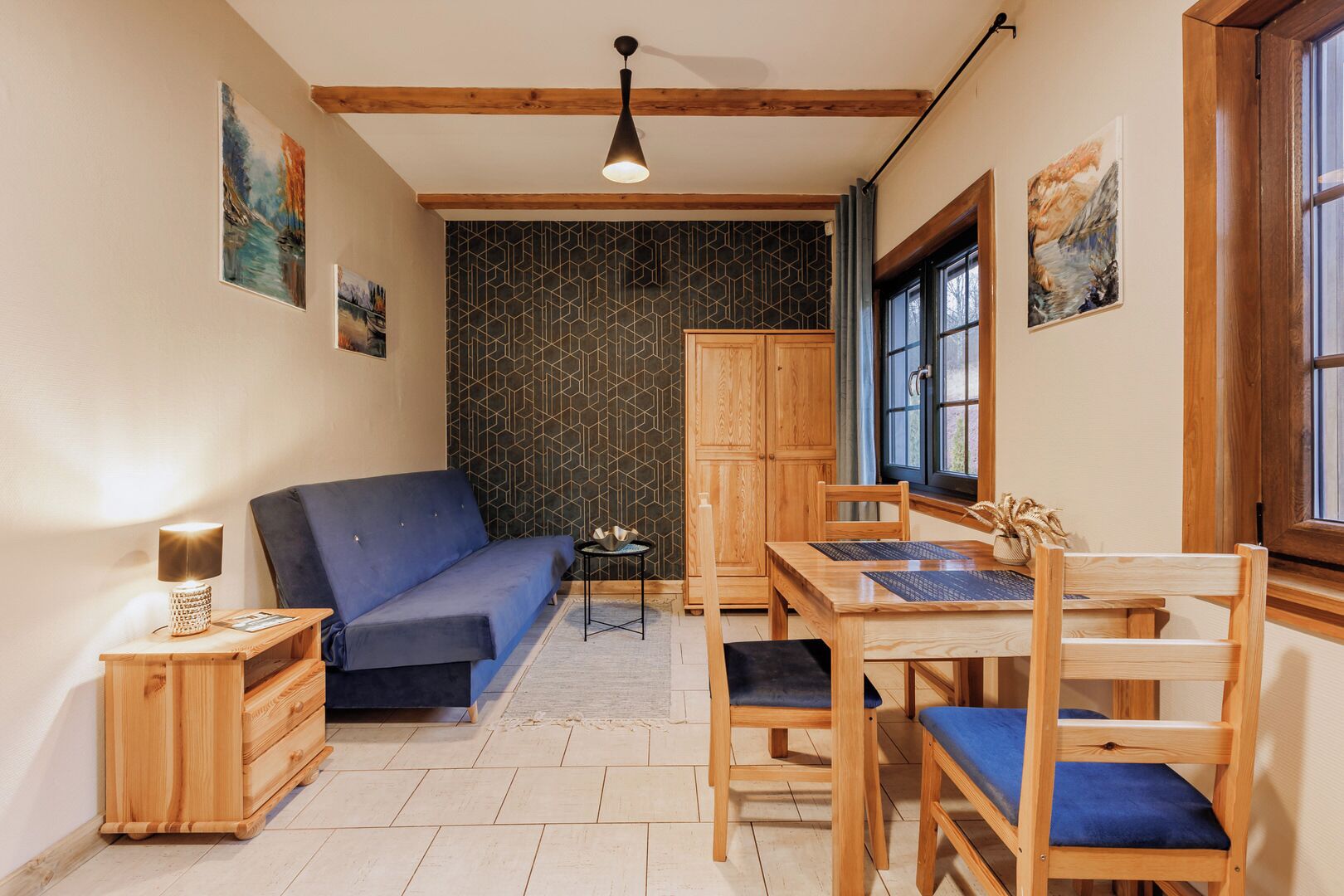 A cozy room with a blue sofa, wooden dining table, and chairs, a small side table with a lamp, and a cabinet.